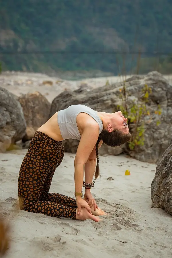 Student holding camel pose on the sand at the riverbank, boulders behind